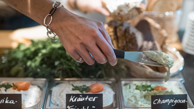 Eine Hand mit Armband nimmt Champignoncreme aus einer Schale. Daneben stehen Schalen mit Kräuterfrischkäse und Paprikacreme, im Hintergrund Brot., © Café Justus, Stuttgart