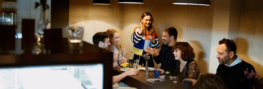A group of people are sitting around a table in a café and laughing. A woman stands and talks to the others. The atmosphere is cozy., © Stuttgart Marketing, Alwin Maigler