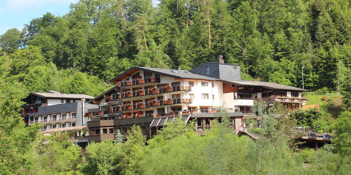 A large hotel in the forest with several balconies, surrounded by lush greenery and trees., © Ringhotel Mönchs Waldhotel