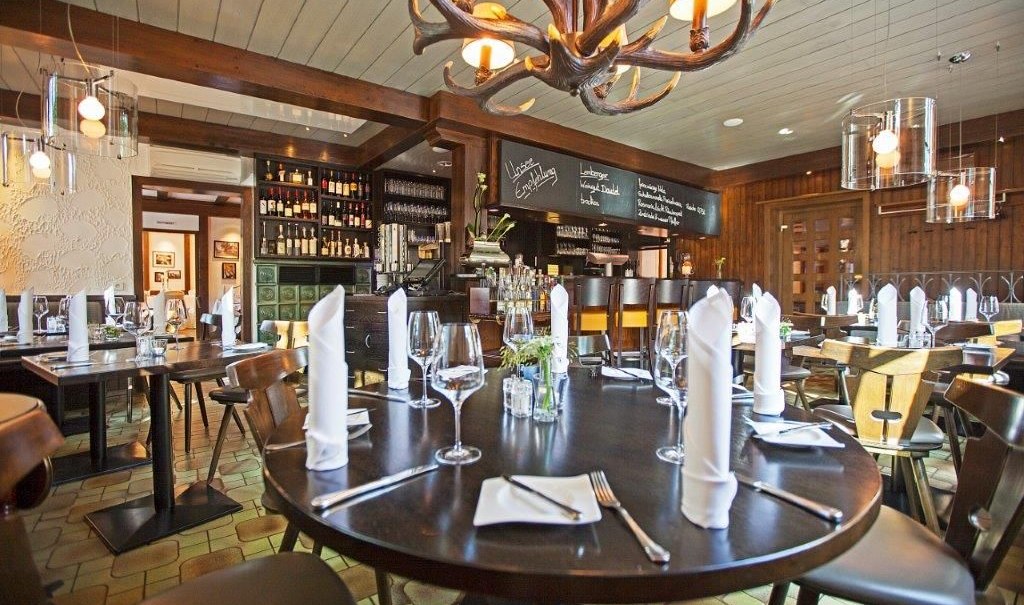 Ein stilvolles Restaurant mit runden Tischen, elegant gedeckt mit Weingläsern und Servietten. Ein Kronleuchter aus Geweihen hängt von der Decke., © Hotel Waldhorn