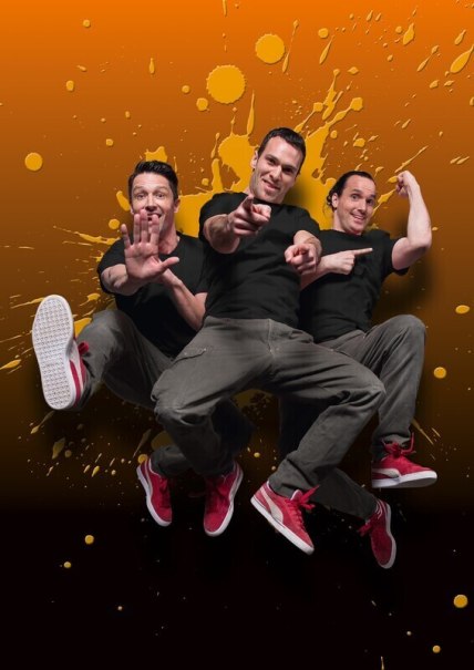 Drei Männer in schwarzen T-Shirts und roten Schuhen springen vor einem orangefarbenen Hintergrund mit Farbspritzern., © Theaterhaus Stuttgart e.V.