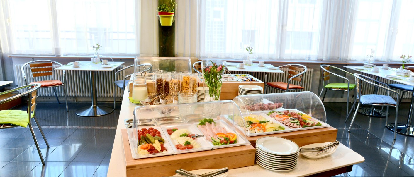 Ein Frühstücksbuffet mit Aufschnitt, Gemüse, Müsli und Geschirr in einem hellen Raum mit Tischen und Stühlen., © Abalon Hotel Ideal Ein Frühstücksbuffet mit Aufschnitt, Gemüse, Müsli und Geschirr in einem hellen Raum mit Tischen und Stühlen., © Abalon Hotel Ideal