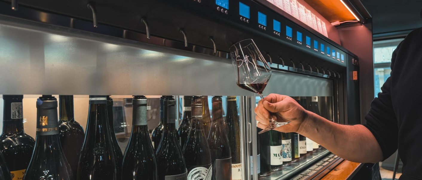 Eine Person hält ein Weinglas mit Rotwein vor einer Reihe von Weinflaschen in einem modernen Weinladen., © Sitt Wein, Stuttgart Eine Person hält ein Weinglas mit Rotwein vor einer Reihe von Weinflaschen in einem modernen Weinladen., © Sitt Wein, Stuttgart