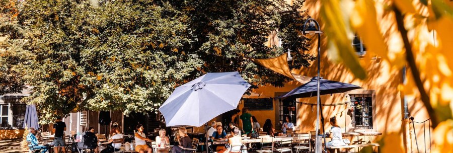 People enjoy the sunny weather in an outdoor caf&eacute;, surrounded by trees and parasols. A relaxed atmosphere prevails., &copy; SMG Stuttgart Marketing GmbH - Sarah Schmid