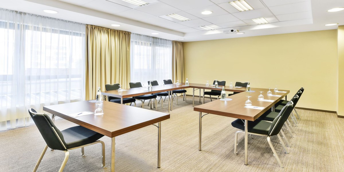 A bright conference room with a U-shaped table arrangement, chairs, water bottles, and glasses. Large windows with curtains., © NH Ludwigsburg A bright conference room with a U-shaped table arrangement, chairs, water bottles, and glasses. Large windows with curtains., © NH Ludwigsburg