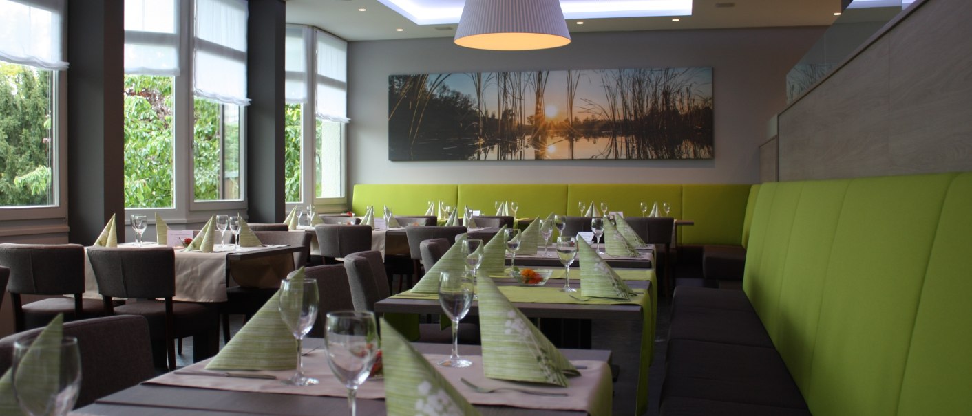 Modernes Restaurant mit gedeckten Tischen, grünen Servietten und großen Fenstern. An der Wand hängt ein Bild mit Sonnenuntergangsmotiv., © Hotel am Schlossberg Herrenberg