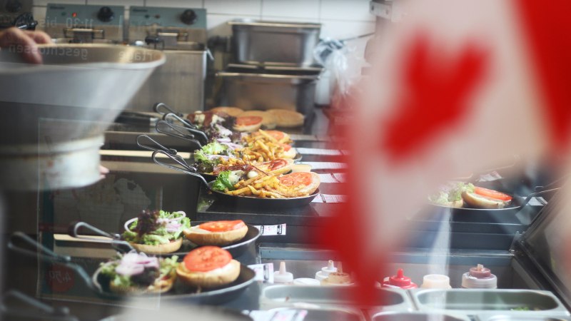 In einer Küche werden Burger und Pommes in Pfannen zubereitet. Im Vordergrund ist unscharf eine kanadische Flagge zu sehen., © SMG