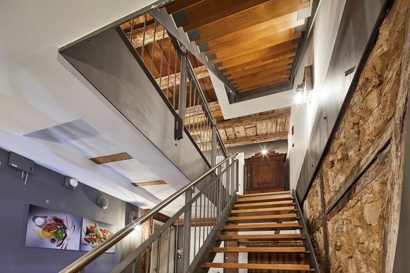 Treppenhaus mit modernen Holzstufen, Metallgel&auml;nder und rustikalen Steinw&auml;nden. An der Wand h&auml;ngen Bilder von Speisen., &copy; Hotel Restaurant Bachofer