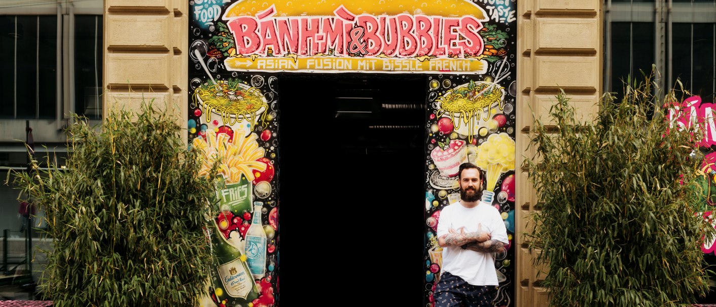 A man stands in front of the entrance to 'Banh Mi & Bubbles', surrounded by colorful graffiti and plants., © Banh Mi & Bubbles Restaurant / Bar