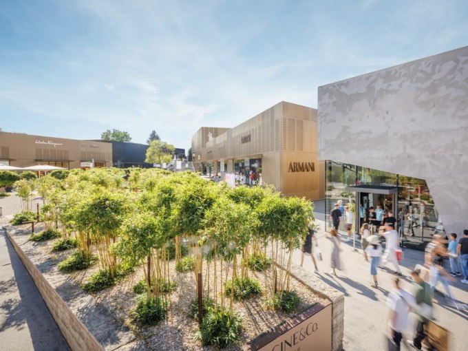 Moderne Einkaufsgebäude in der Outletcity Metzingen mit Menschen, die entlang der Geschäfte flanieren. Grüne Pflanzen im Vordergrund., © Schlienz-Tours GmbH & Co.KG Moderne Einkaufsgebäude in der Outletcity Metzingen mit Menschen, die entlang der Geschäfte flanieren. Grüne Pflanzen im Vordergrund., © Schlienz-Tours GmbH & Co.KG
