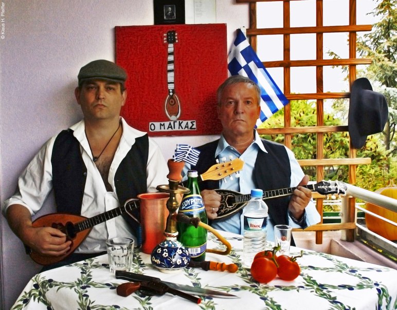 Zwei Männer in traditioneller Kleidung spielen griechische Instrumente an einem Tisch mit Getränken, Tomaten und einer griechischen Flagge im Hintergrund., © Fotocredit Theater La Lune/LEFTA rembetiko