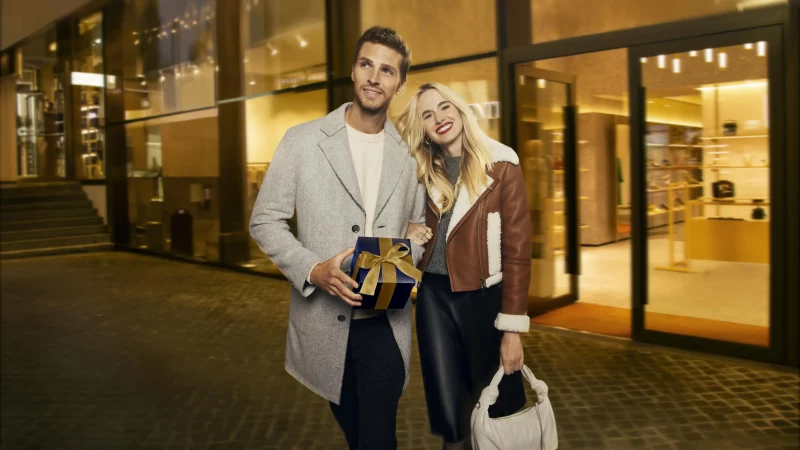 A smiling couple in front of a lit-up store; the man is holding a gift box, and the woman is carrying a handbag. Festive and elegant., © OUTLETCITY AG A smiling couple in front of a lit-up store; the man is holding a gift box, and the woman is carrying a handbag. Festive and elegant., © OUTLETCITY AG