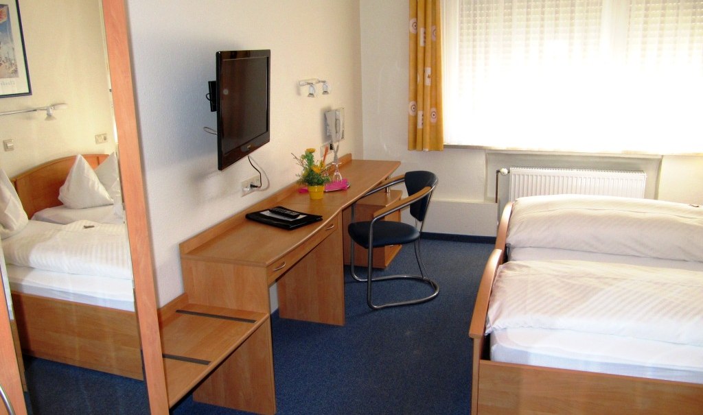 Ein schlichtes Gästezimmer mit zwei Einzelbetten, einem Schreibtisch mit Stuhl, einem Fernseher an der Wand und einem Fenster mit gelben Vorhängen., © R.Storr Ein schlichtes Gästezimmer mit zwei Einzelbetten, einem Schreibtisch mit Stuhl, einem Fernseher an der Wand und einem Fenster mit gelben Vorhängen., © R.Storr