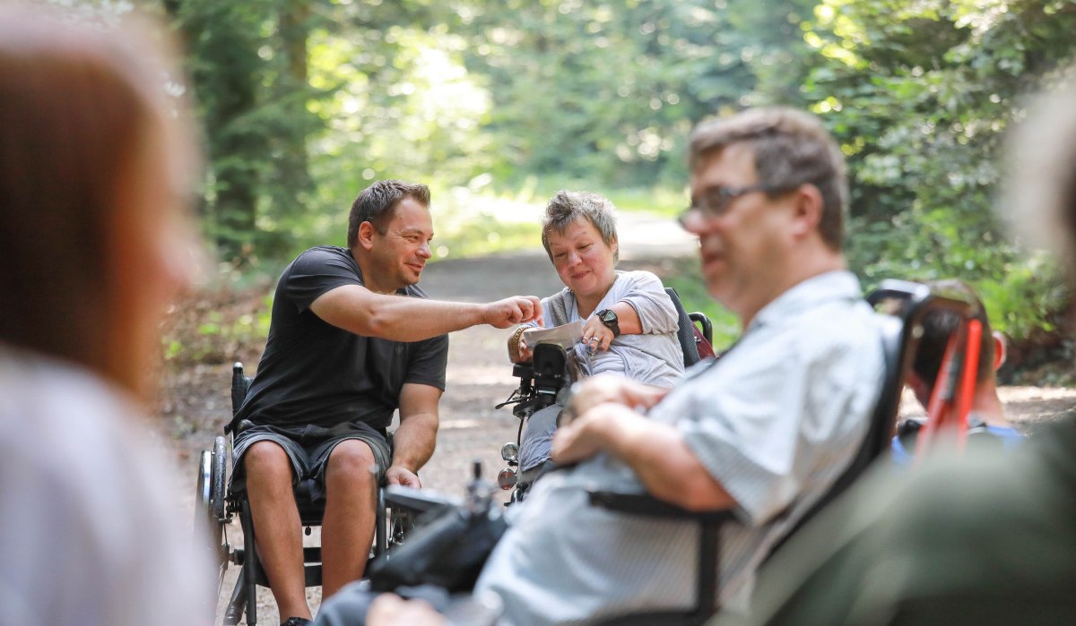 Eine Gruppe von Menschen in Rollst&uuml;hlen unterh&auml;lt sich auf einem Waldweg. Die Umgebung ist gr&uuml;n und sonnig., &copy; NPSFW