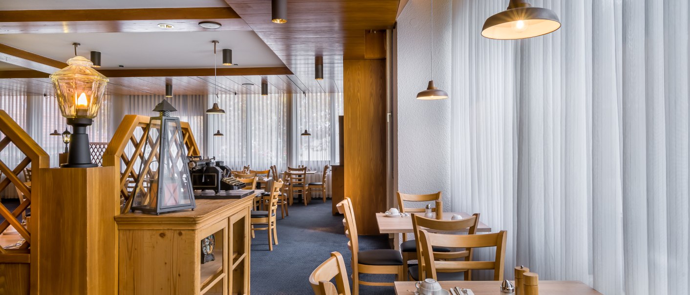 Ein Frühstücksbereich mit Holzmöbeln, gedeckten Tischen und warmem Licht. Große Fenster mit Vorhängen lassen Tageslicht herein., © Novum Management GmbH Ein Frühstücksbereich mit Holzmöbeln, gedeckten Tischen und warmem Licht. Große Fenster mit Vorhängen lassen Tageslicht herein., © Novum Management GmbH