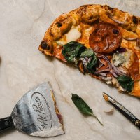 Halb gegessene Pizza mit Basilikum und Zwiebeln auf hellem Stein, daneben Pizzaschneider und Schere; links zwei t&auml;towierte H&auml;nde., &copy; Connective Media