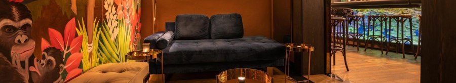 Gemütliche Lounge mit blauer Couch, buntem Wandbild mit Affen und Pflanzen. Im Hintergrund eine Bar mit beleuchteten Regalen und Hockern., © Holzmaler, Stuttgart