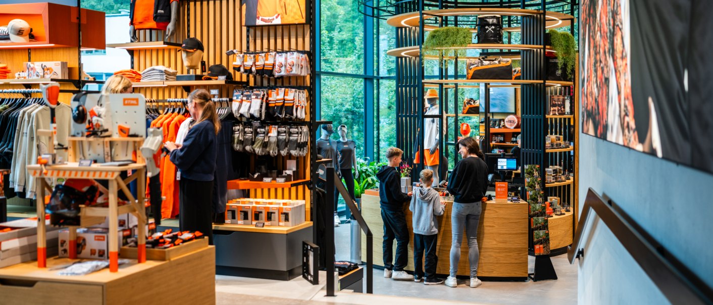 A STIHL brand store featuring clothing, accessories, and customers at the register. Modern interior design with wooden shelves and large windows., © STIHL Markenwelt A STIHL brand store featuring clothing, accessories, and customers at the register. Modern interior design with wooden shelves and large windows., © STIHL Markenwelt