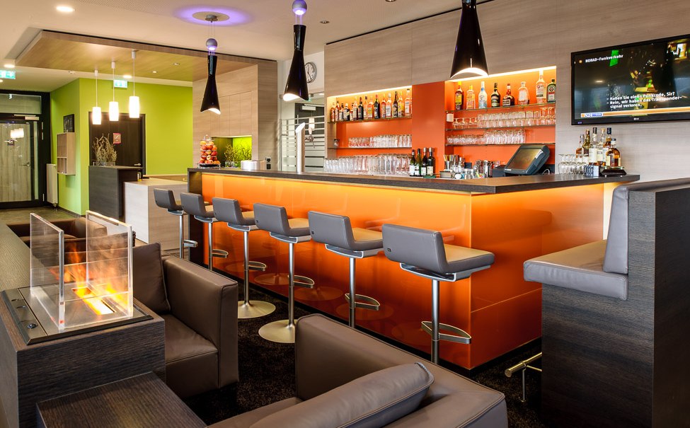 Moderne Bar mit orange beleuchtetem Tresen, grauen Barhockern und Flaschenregal. Im Vordergrund gemütliche Sitzgelegenheiten und ein kleiner Kamin., © TOMAS Moderne Bar mit orange beleuchtetem Tresen, grauen Barhockern und Flaschenregal. Im Vordergrund gemütliche Sitzgelegenheiten und ein kleiner Kamin., © TOMAS