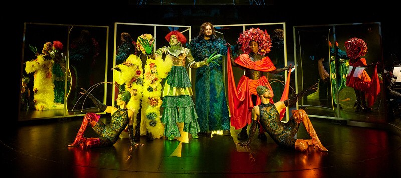 Bühnenbild mit Darstellern in bunten, fantasievollen Kostümen vor Spiegeln. Die Szene wirkt lebendig und kreativ inszeniert., © Württembergische Staatstheater Stuttgart