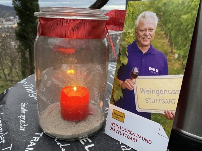Ein Glas mit rotem Band und Kerze steht neben einem Flyer für Weintouren in Stuttgart. Im Hintergrund sind Weinberge zu sehen., © Weingenuss Stuttgart Ein Glas mit rotem Band und Kerze steht neben einem Flyer für Weintouren in Stuttgart. Im Hintergrund sind Weinberge zu sehen., © Weingenuss Stuttgart