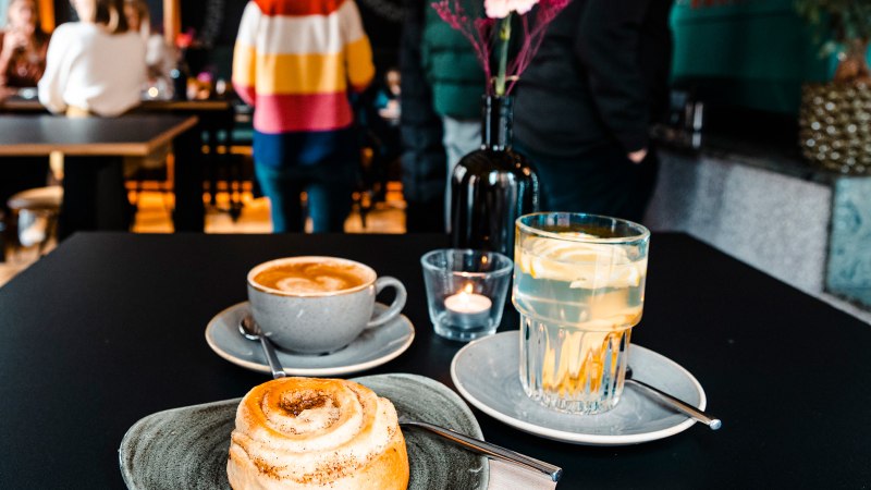 Ein Tisch in einem Café mit Zimtschnecke, Cappuccino und Zitronenwasser. Im Hintergrund sind unscharfe Personen zu sehen., © Stuttgart-Marketing GmbH, Sarah Schmid