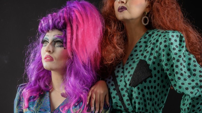 Zwei Drag Queens in bunten, auffälligen Outfits und extravagantem Make-up posieren selbstbewusst vor einem dunklen Hintergrund., © Veranstalter Zwei Drag Queens in bunten, auffälligen Outfits und extravagantem Make-up posieren selbstbewusst vor einem dunklen Hintergrund., © Veranstalter