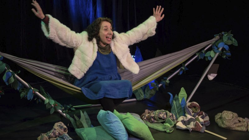 Frau in weißer Jacke und blauem Kleid sitzt in Hängematte, Arme ausgestreckt. Umgeben von grünen Kissen und Decken auf einer Bühne., © Àngels Capell