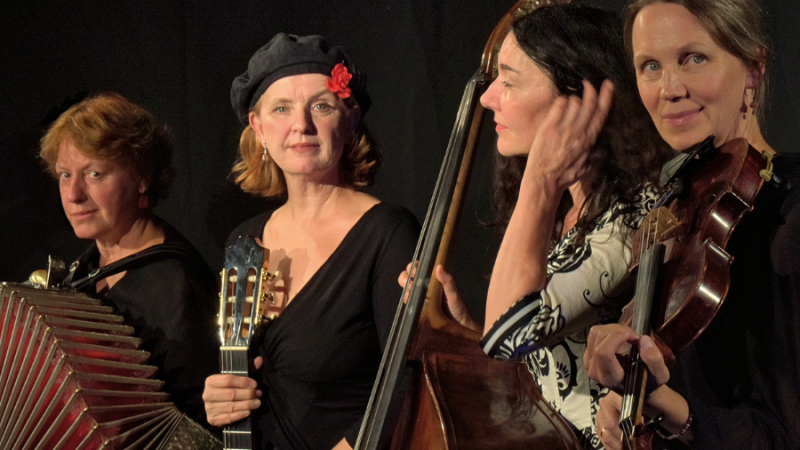 Four musicians with accordion, guitar, double bass and violin pose in front of a dark background. One woman is wearing a black beret with a red flower., &copy; Fotocredit Theater La Lune Stuttgart