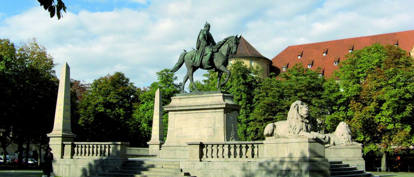 Equestrian statue on Karlsplatz in Stuttgart, flanked by lion statues and obelisks, surrounded by trees and a historic building in the background., © Stuttgart-Marketing GmbH