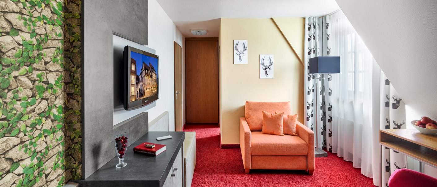 Ein gemütliches Einzelzimmer mit orangenem Sessel, rotem Teppich, TV und Wanddekoration mit Hirschmotiven. Ein Fenster mit Vorhängen lässt Licht herein., © Hirsch Hotel