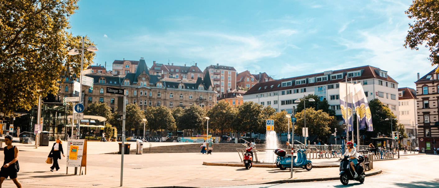 Städtischer Platz mit Springbrunnen, umgeben von historischen Gebäuden. Menschen gehen und fahren Roller bei sonnigem Wetter. Schilder und Bäume im Hintergrund., © SMG Stuttgart Marketing GmbH - Sarah Schmid Städtischer Platz mit Springbrunnen, umgeben von historischen Gebäuden. Menschen gehen und fahren Roller bei sonnigem Wetter. Schilder und Bäume im Hintergrund., © SMG Stuttgart Marketing GmbH - Sarah Schmid