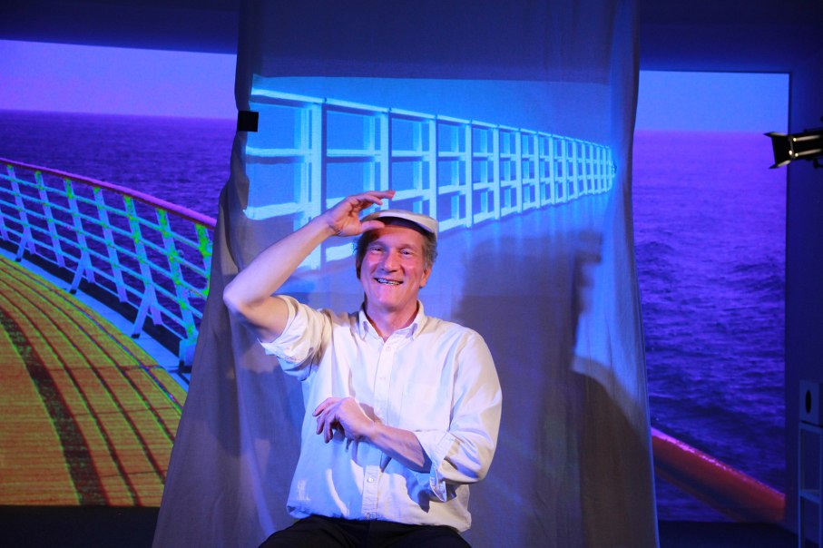 A man in a white shirt and hat sits smiling in front of a screen showing the deck of a ship. The scene is reminiscent of a theater performance., &copy; Katja Ritter