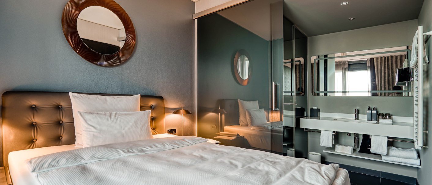 Modernes Hotelzimmer mit einem großen Bett, einem runden Spiegel an der Wand und einem angrenzenden Badezimmer mit Glaswand., © CLOUD N°7 APARTMENTS GmbH