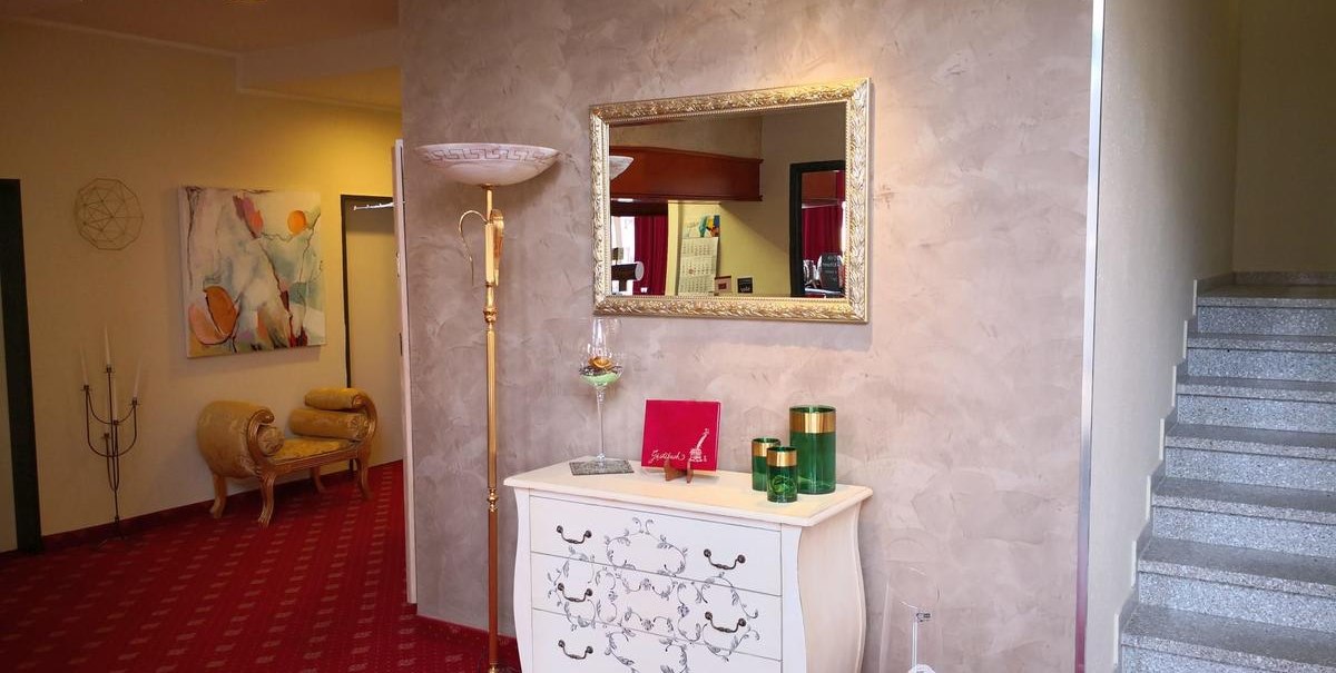 Elegante Lobby mit rotem Teppich, einer weißen Kommode, Spiegel, Stehlampe und dekorativen Elementen. Treppe rechts, Gemälde links., © Hotel Arcis
