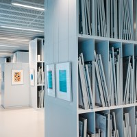 Interior view of a modern art library with shelves full of framed works of art and light-colored walls. Pictures hang on the walls., © Sarah Schmid