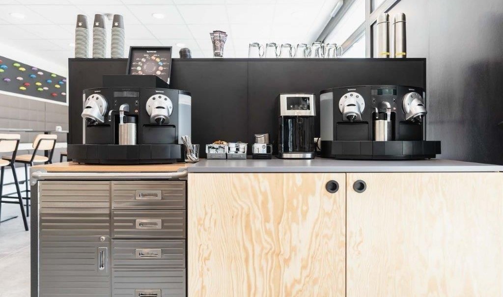 Moderne Kaffestation mit zwei Kaffeemaschinen, Bechern und Zubehör auf einem Holzschrank in einem hellen Raum., © a2 HOTELS am Quadrium