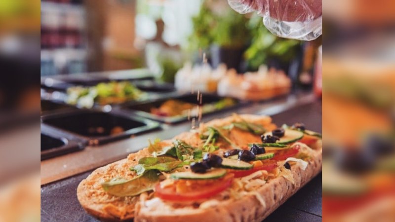 A sandwich is topped with fresh vegetables and olives. A hand sprinkles spices on top. Ingredients in containers can be seen in the background., &copy; FreshSub