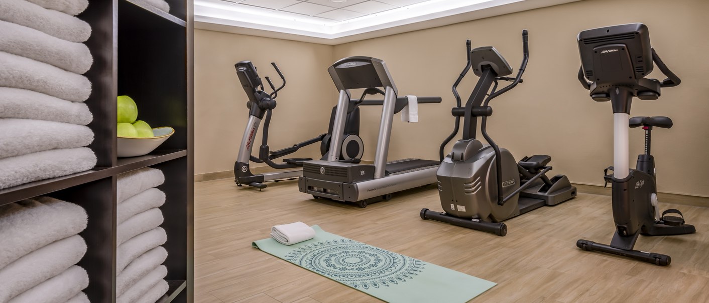 Fitness room with treadmill, cross trainer, exercise bike, yoga mat and towels. A shelf with towels and apples stands in the foreground., © Stuttgart Marriott Hotel Sindelfingen Fitness room with treadmill, cross trainer, exercise bike, yoga mat and towels. A shelf with towels and apples stands in the foreground., © Stuttgart Marriott Hotel Sindelfingen
