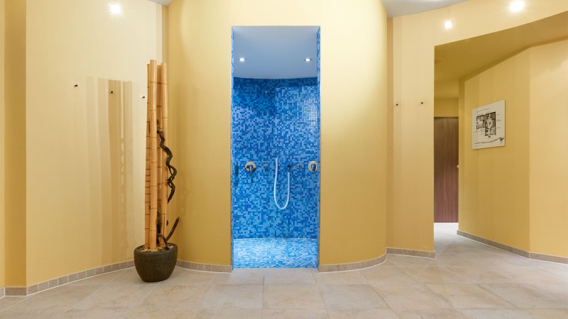 Ein Spa-Bereich mit einer blauen Mosaikdusche in der Mitte, umgeben von gelben Wänden und dekorativen Bambusstäben., © TOMAS Ein Spa-Bereich mit einer blauen Mosaikdusche in der Mitte, umgeben von gelben Wänden und dekorativen Bambusstäben., © TOMAS