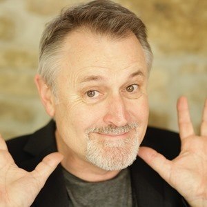 A man with gray hair and beard smiles and raises his hands in a cheerful gesture against a blurred background., &copy; Renitenztheater Stuttgart e.V.