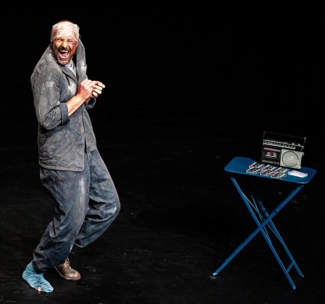 Ein Mann in staubiger Kleidung steht auf einer B&uuml;hne und wirkt begeistert. Neben ihm steht ein Tisch mit einem Radio und mehreren Kassetten., &copy; Theaterhaus Stuttgart e.V.