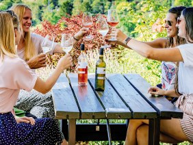 Fünf Personen stoßen mit Weingläsern an einem Holztisch im Freien an, umgeben von grüner Natur und Blumen., © SMG, Thomas Niedermüller Fünf Personen stoßen mit Weingläsern an einem Holztisch im Freien an, umgeben von grüner Natur und Blumen., © SMG, Thomas Niedermüller