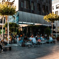Menschen genießen das sonnige Wetter in einem belebten Außenbereich eines Cafés. Moderne Gebäude und Bäume umgeben die Szene., © SMG, Sarah Schmid
