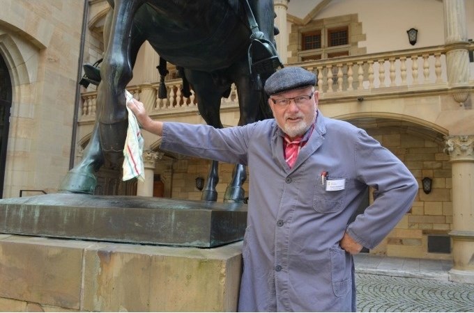 Ein Mann in blauer Arbeitskleidung und Mütze steht neben einer Pferdestatue und hält ein Tuch. Er befindet sich in einem historischen Innenhof., © Stuttgart Marketing GmbH