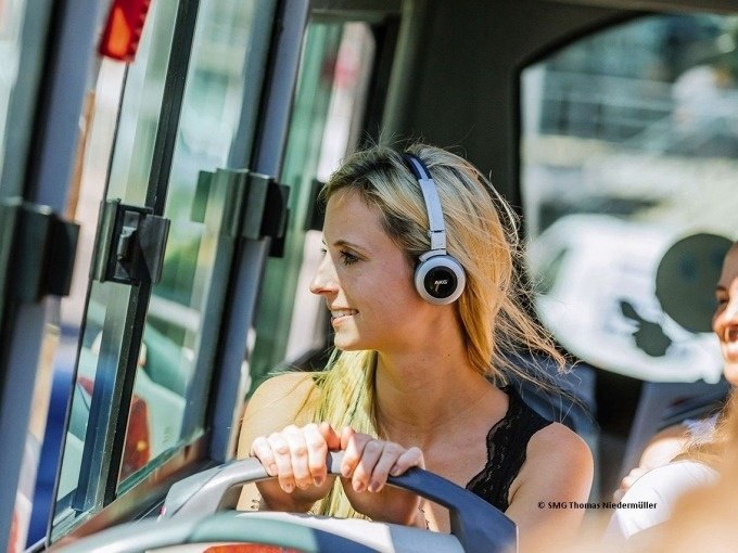 Eine Frau mit Kopfh&ouml;rern sitzt in einem Bus und schaut l&auml;chelnd aus dem Fenster. Sonnenlicht f&auml;llt durch die Scheiben., &copy; SMG, Thomas Niederm&uuml;ller