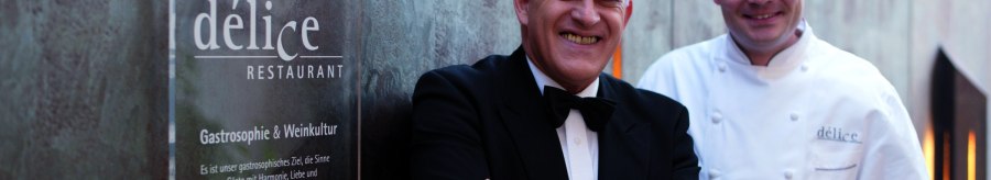 Zwei M&auml;nner stehen vor einem Schild des D&eacute;lice Restaurants. Einer tr&auml;gt einen Anzug, der andere eine Kochjacke., &copy; Delice