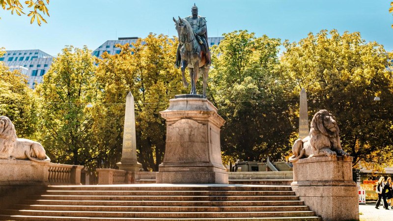 Reiterstatue auf dem Karlsplatz, flankiert von zwei Löwenstatuen. Umgeben von Bäumen und modernen Gebäuden im Hintergrund., © Stuttgart Marketing GmbH, Sarah Schmid Reiterstatue auf dem Karlsplatz, flankiert von zwei Löwenstatuen. Umgeben von Bäumen und modernen Gebäuden im Hintergrund., © Stuttgart Marketing GmbH, Sarah Schmid