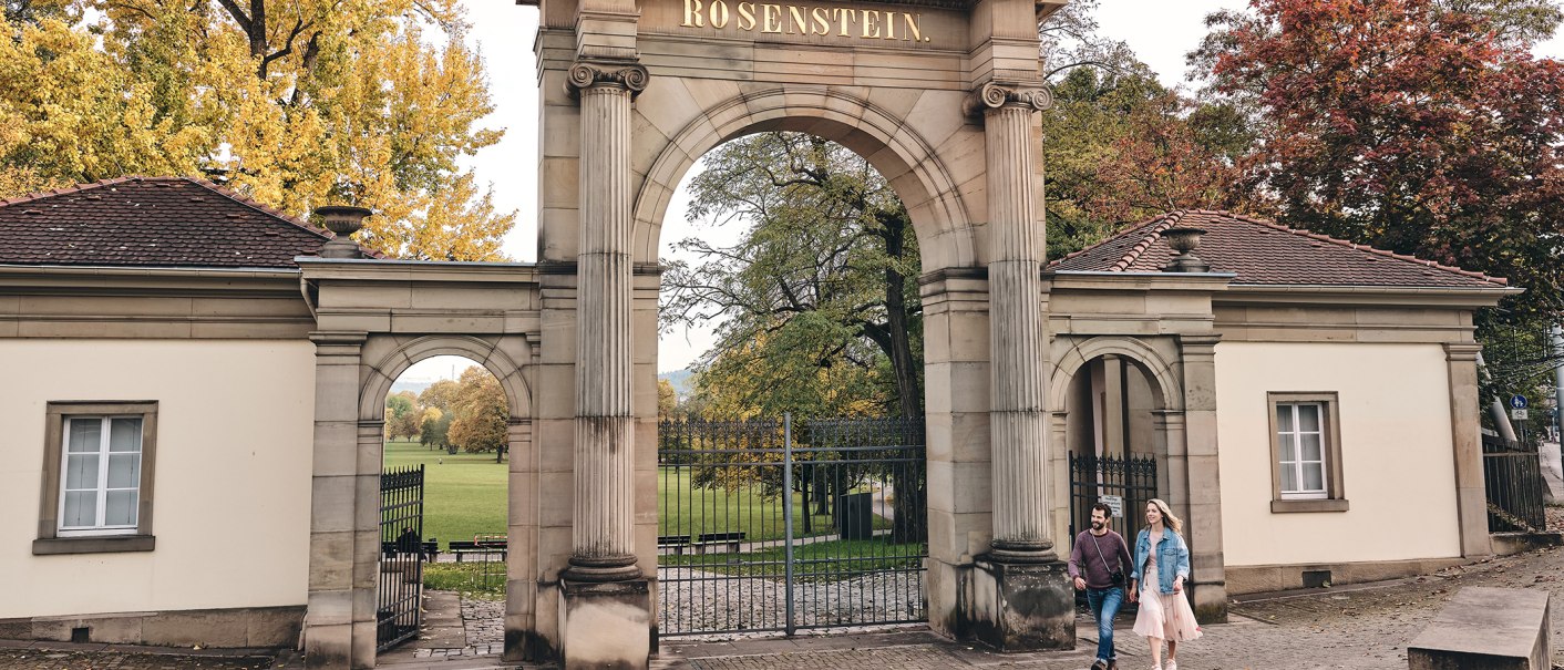 Entrance gate to Rosenstein Park with the inscription 'Rosenstein'. Two people walk past, surrounded by autumnal trees., © SMG, Christoph Düpper Entrance gate to Rosenstein Park with the inscription 'Rosenstein'. Two people walk past, surrounded by autumnal trees., © SMG, Christoph Düpper