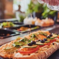 A sandwich is topped with fresh vegetables and olives. A hand sprinkles spices on top. Ingredients in containers can be seen in the background., &copy; FreshSub